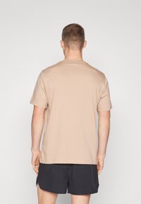 Beige T-Shirt mit kurzen Ärmeln und Rundhalsausschnitt, glatte Textur und lockere Passform, kombiniert mit dunklen Shorts, von hinten sichtbar.