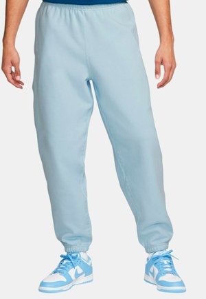 Light blue jogger pants made of a smooth fabric, featuring an elastic waistband, tapered ankles, and side seam pockets, paired with blue sneakers.