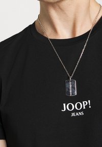 Černé tričko s bílým nápisem "JOOP! JEANS", s stříbrným řetízkem a kovovým přívěškem ve tvaru psí známky s texturovaným povrchem.