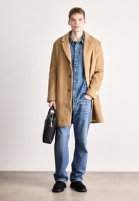 Παλιό παλτό από καμήλα πάνω από πουκάμισο τζην, συνδυασμένο με μπλε τζιν και μαύρες σουέτ μπότες. Το μοντέλο κρατά μια μαύρη δερμάτινη τσάντα.