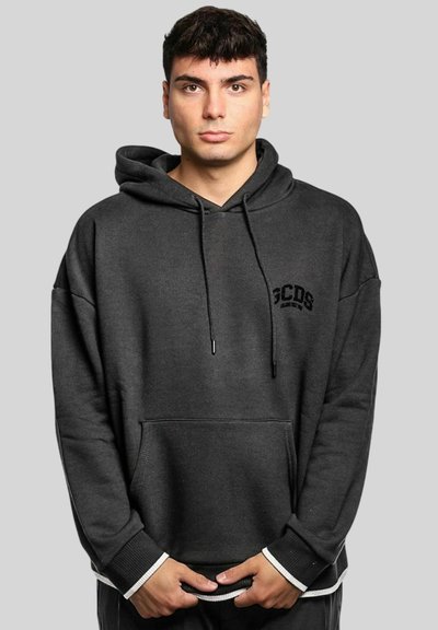 Jeune homme aux cheveux foncés portant un sweat à capuche noir GCDS, debout devant un fond gris clair uni, les mains jointes devant lui.