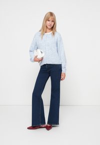 Jasnoniebieski, dziany kardigan z guzikami, w połączeniu z ciemnoniebieskimi, rozszerzającymi się dżinsami i czerwonymi płaskimi butami. Trzyma małą białą torebkę.