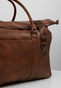 Braune Leder-Duffel-Tasche mit strukturiertem Oberflächen, Reißverschluss, Stoffgurt und Metallbeschlägen. Verfügt über eine Seitentasche mit eingeprägtem Logo.