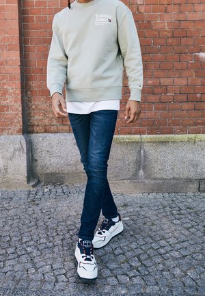 Osoba ubrana w jasnoszarą bluzę Tommy Jeans, ciemnoniebieskie jeansy i białe trampki, idąca po bruku obok czerwonej ceglanej ściany.