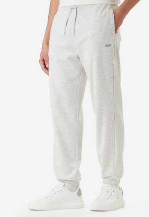 Uomo che indossa pantaloni della tuta grigio chiaro con coulisse in vita e sneakers bianche, in piedi contro uno sfondo semplice.
