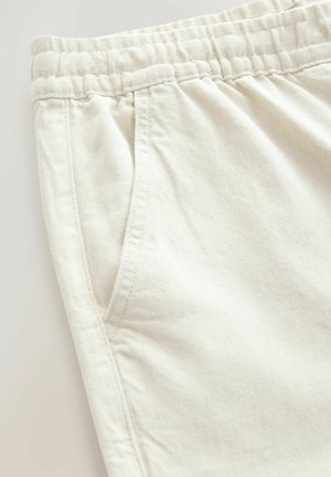 Pantalons en coton beige clair avec une taille élastique, des poches latérales et une texture diagonale fine. Les coutures sont visibles près des poches.