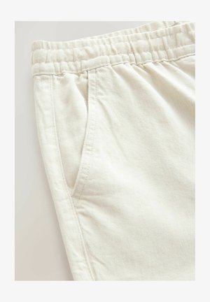 Pantaloni din bumbac bej deschis, cu un band elastic la talie, buzunare laterale și o textură fină în diagonală. Detalii de cusătură sunt vizibile lângă buzunare.