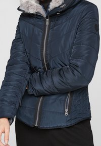 Osoba v námořnickém prošívaném kabátu s kapucí osvětlenou kožešinou a se zipovými kapsami, s překříženýma rukama na hrudi, na jednobarevném pozadí.