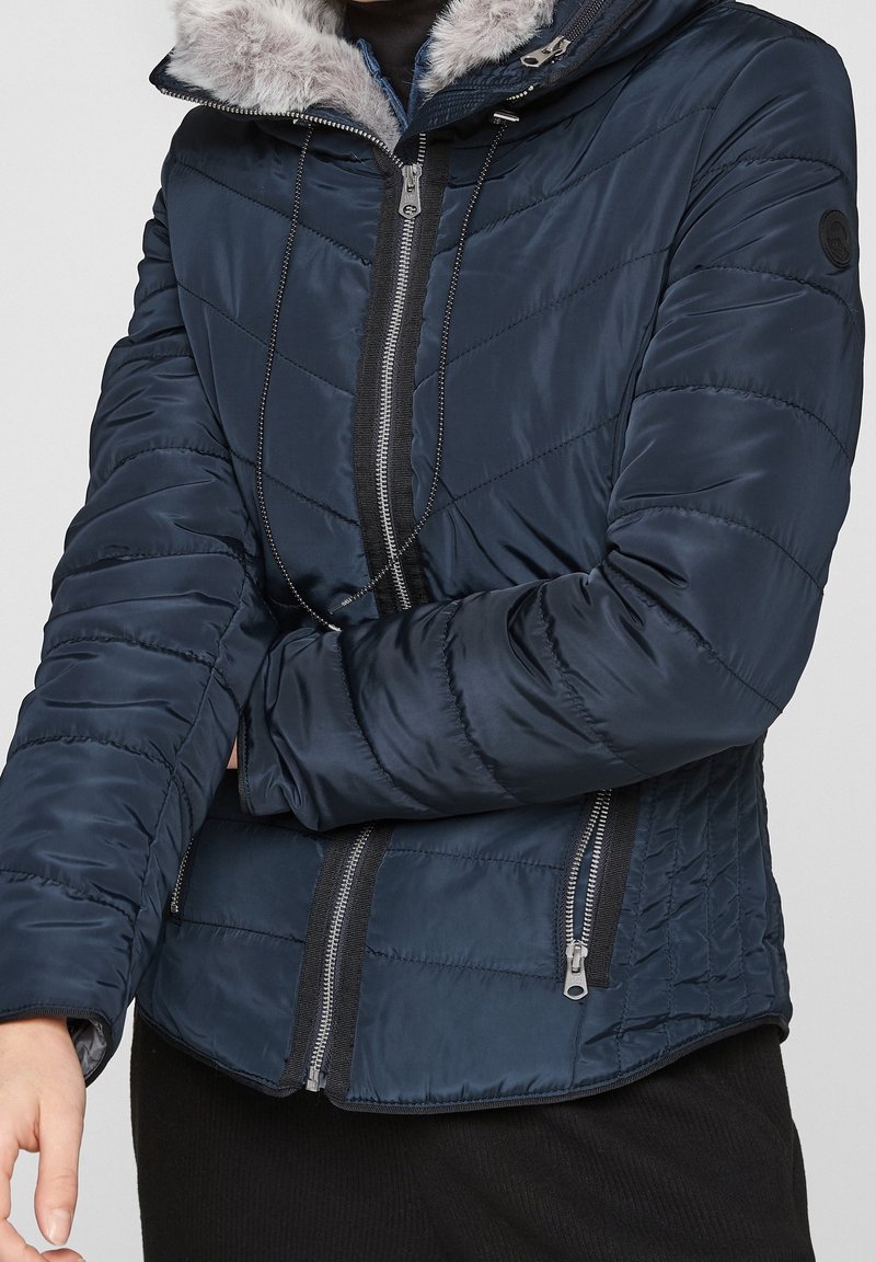 Osoba v námořnickém prošívaném kabátu s kapucí osvětlenou kožešinou a se zipovými kapsami, s překříženýma rukama na hrudi, na jednobarevném pozadí.