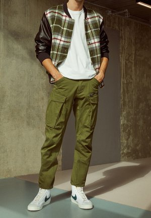 Osoba ubrana w zielone spodnie bojówki, białe trampki, białą koszulkę i zieloną kurtkę w kratę z czarnymi skórzanymi rękawami, stojąca w pomieszczeniu.