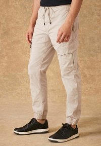 Pantaloni cargo beige con polsini elasticizzati, dotati di tasche laterali e una vita con cordino, abbinati a sneakers nere. Sfondo testurizzato.