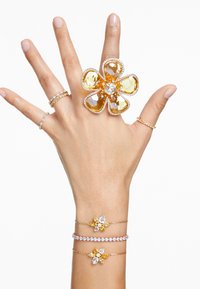 Grande anel de flor amarela com pedras preciosas claras e amarelas, acompanhado por duas pulseiras delicadas com realces em amarelo e claro, tudo em tom dourado.
