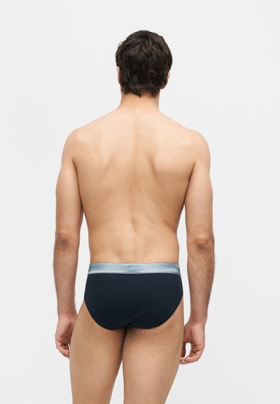 Άνδρας με κοντά σκούρα μαλλιά που στέκεται γυρισμένος από πίσω, φοράει σκούρα εσώρουχα με ανοιχτό γκρι ζώνη στη μέση, σε απλό ανοιχτό φόντο.