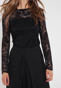 Top in pizzo nero a maniche lunghe con scollatura a onda e complessi motivi floreali, abbinato a una gonna nera a vita alta.