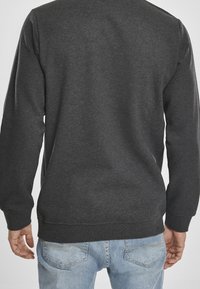 Uomo che indossa una felpa grigia scura a maniche lunghe e jeans azzurri chiari, visto da dietro su uno sfondo semplice.