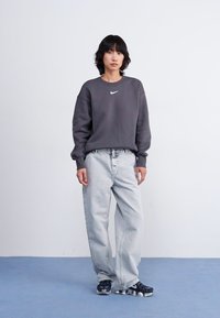 Grijze sweatshirt met een klein wit logo, gecombineerd met lichtblauwe baggy jeans en zwarte sportschoenen met een gestructureerde zool. Eenvoudige achtergrond.
