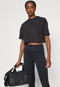 Μαύρο κοντομάνικο τοπ από μαλακό υλικό, σε συνδυασμό με μαύρο κολάν ψηλής μέσης. Το μο ντέλο κρατάει μια μαύρη τσάντα τύπου duffel με ανθεκτικούς ιμάντες.