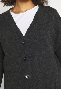 Kolsvart cardigan i texturerat tyg, med V-hals, fyra svarta knappar och lager över en vit skjorta.