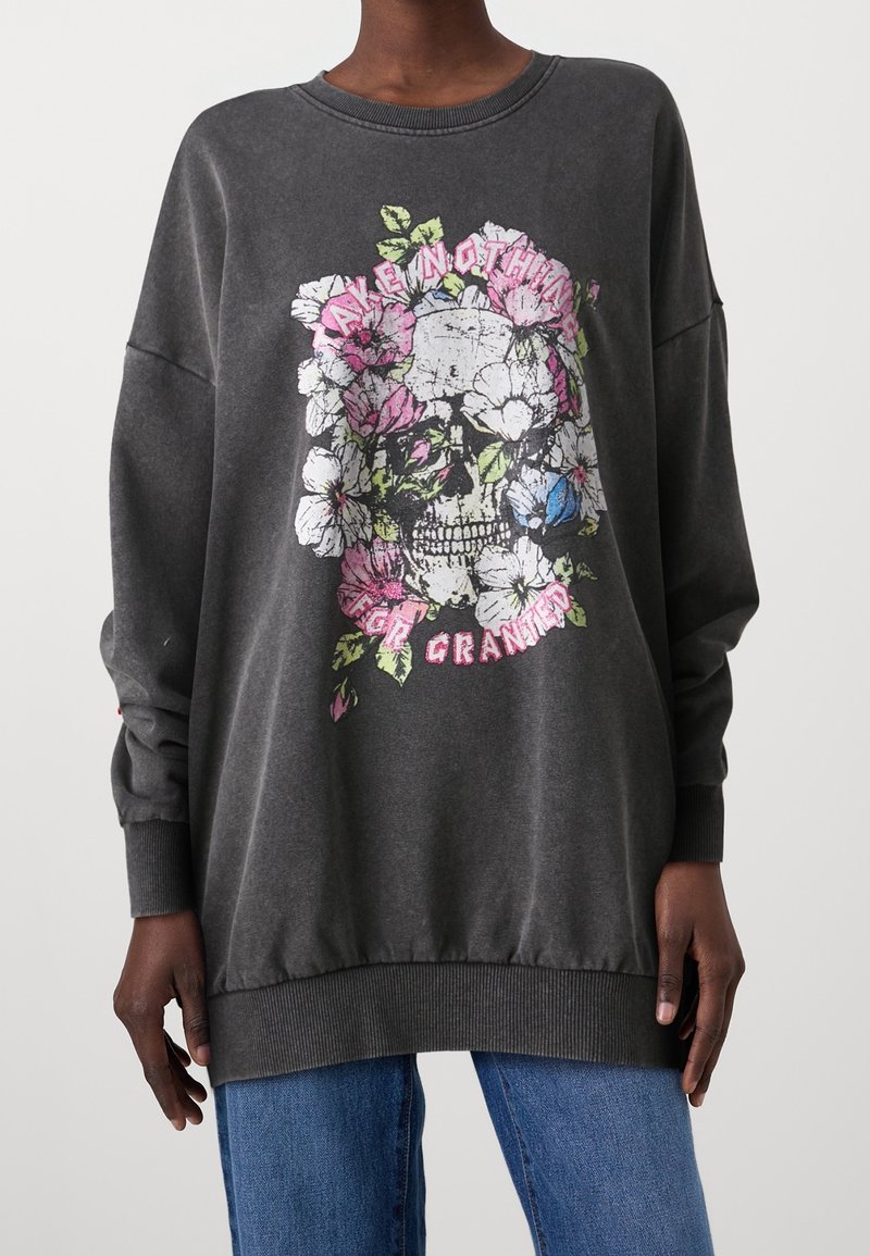 Personne portant un sweat-shirt oversize gris foncé avec un crâne entouré de fleurs roses et blanches et un texte rose à l'avant.