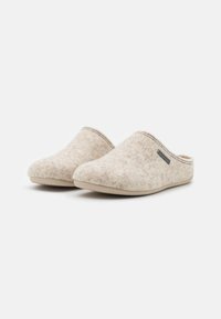 Chaussons en feutre doux de couleur beige clair, avec une texture lisse, un design à l'arrière fermé et une semelle en caoutchouc beige. Comprend un petit logo en accent.