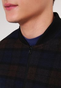 Ala al-ujuh wa al-‘uqā' li-shakhs yalbas jaqit za'i al-mashaud miti'a ‘ala qamīṣ azraq, ma'a sha‘ar wajh khafī ‘ala al-daqn.