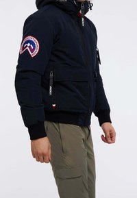 Veste bomber bleu marine avec capuche bordée de fourrure, poches zippées et écusson brodé sur la manche gauche. Détails noirs et rouges en accents.
