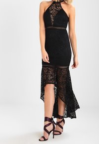 Robe noire en dentelle avec un bustier ajusté et un ourlet à volant, présentant des motifs complexes et des accents transparents. Associée à des talons à brides.