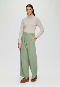 Hellbeige Rollkragenoberteil, hoch taillierte, gefaltete grüne Hose und braune Mokassins. Glatte Stoffoberfläche mit angepasster Passform und geradem Schnitt.
