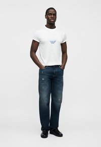 Muškarac stoji s rukama u džepovima, noseći bijelu majicu s grafičkim uzorkom, tamnoplave izlizane traperice i crne tenisice, na jednostavnoj pozadini.