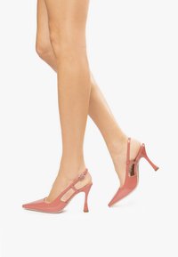 Różowe, lakierowane czółenka na obcasie slingback z wąskimi noskami, zwężającymi się obcasami i regulowanymi paskami na kostkę. Zawierają wycięcia po obu stronach.