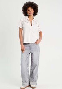 Blouse blanche à manches courtes avec des détails en broderie, associée à un jean large gris clair. La tenue présente une coupe décontractée et un design à bouton.