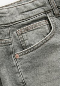 Jasnoszary materiał dżinsowy z bliska, widoczne szwy, kieszeń boczna oraz miedziany nit na pasku.