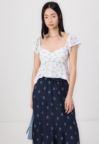 Blusa bianca floreale con maniche corte a sbuffo, abbinata a una gonna a strati blu scuro con motivo a foglie. Design casual, tessuto leggero.