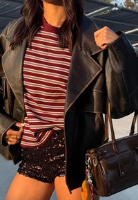 Svart läderjacka över en vinröd randig skjorta, i kombination med svarta paljettshorts och en brun handväska. Texturerade material och lyster synliga.