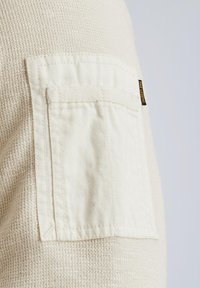 Tissu texturé de couleur crème avec une poche sur la poitrine gauche en matériau blanc lisse. Présente une étiquette sur la poche.