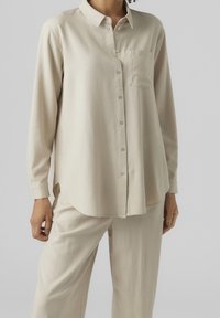 Beige Langarm-Hemd mit Knöpfen, Kragen und einer Brusttasche. Weicher Stoff, lockere Passform, abgerundeter Saum und einfache Knopfverschluss.