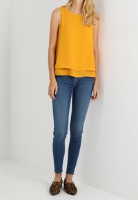 Geel mouwloos topje met gelaagd ontwerp, gecombineerd met blauwe skinny jeans en bruine loafers met panterprint. Neutrale achtergrond.