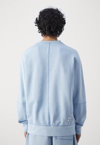 Person med lockigt mörkt hår som bär en ljusblå sweatshirt och matchande byxor, vänd bort från en enkel vit bakgrund.