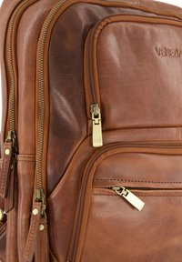 Hnědý kožený batoh s několika zipovými kapsami a mosazným kováním. Hladký povrch s viditelnými švy a detailem s logem.