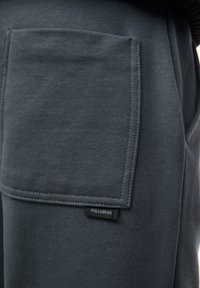 Mörkgrå bomullsjoggers med en stor ficka och en liten "PULL&BEAR"-etikett nära fållen. Slät textur och avslappnad design.