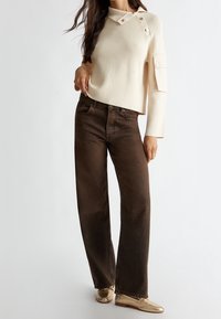 Pull en maille côtelée crème avec boutons en accent, associé à un jean marron à jambes larges. Le mannequin porte des chaussures métalliques dorées. Fond simple.