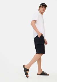 Witte V-hals T-shirt, marineblauwe cargoshorts met zijzakken, en zwarte sandalen met twee verstelbare banden. Eenvoudig ontwerp; gladde textuur.