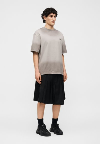 Νέος άνδρας με σγουρά μαλλιά, που φοράει χαλαρό γκρι t-shirt, μαύρη πλισέ φούστα μέχρι το γόνατο, μαύρες κάλτσες και μαύρα αθλητικά παπούτσια, στέκεται μπροστά σε λευκό φόντο.