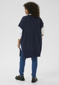 Cardigan lung de culoare navy din tricot, cu mâneci scurte, fante laterale și un siluetă dreaptă, purtat peste o cămașă albă și asortat cu blugi albaștri.