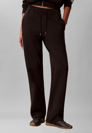 Osoba w czarnych spodniach dresowych ze ściągaczem, z białymi paskami po bokach, oraz czarnych butach sportowych z gumową podeszwą, stojąca na szarym tle.