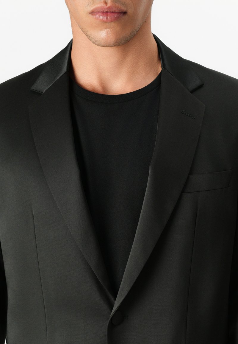 Blazer noir avec revers en satin, coupe ajustée, fermeture à un bouton et un détail de poche subtil. Porté par-dessus un t-shirt noir à col rond.