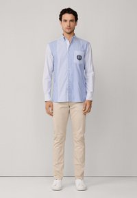 Gestreiftes blau-weißes Hemd mit Button-Down-Kragen, einer einzelnen Brusttasche und Logo-Patch, kombiniert mit beigen Hosen und weißen Sneakers.