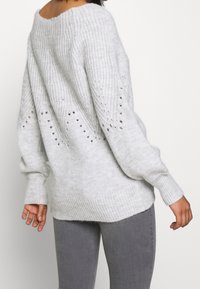 Pull en tricot gris clair avec des détails en tissage ouvert, col large et poignets côtelés. Texture douce avec une coupe décontractée et un dos plus long.