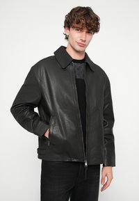 Veste en cuir noire avec un large col, fermeture éclair à l'avant et poches latérales. Texture lisse et design décontracté, portée sur un haut noir.