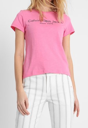 Pink short-sleeved t-shirt with round neckline, featuring "Calvin Klein Jeans" text in black. Paired with white vertical-striped pants.